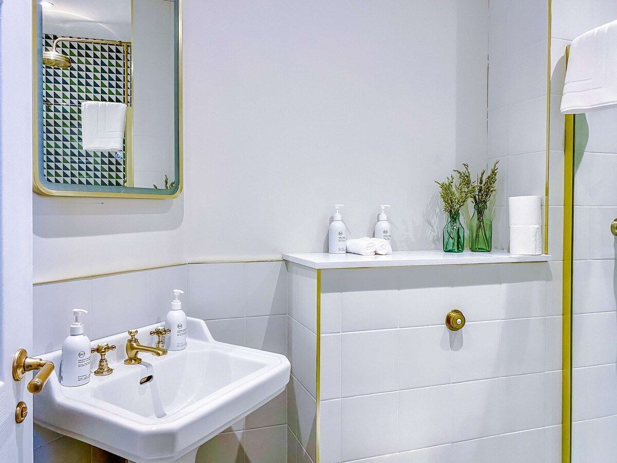 A bathroom with a white sink, gold faucets, and a mirror. There are toiletries on the counter and a towel hanging on the wall.
