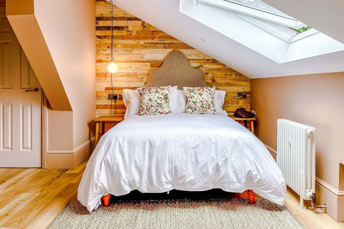 A bedroom with a sloped ceiling, a skylight, a bed with white bedding, and a wooden wall.