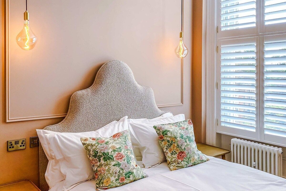 A hotel room with a bed, two bedside lamps, a radiator, and a window with blinds.