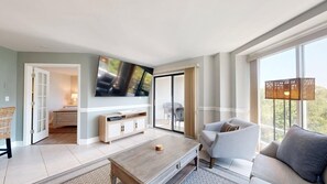 A modern living room with a flat-screen TV mounted on the wall, a sofa, a coffee table, and a view of the outdoors through large windows. - 2 Bedroom close to the beach! (Hilton Head)