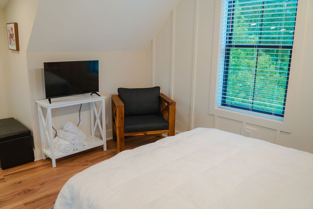 Ein Schlafzimmer mit einem Bett, einem Fernseher auf einem weißen Schrank, einem Holzstuhl und einem Fenster mit Jalousien.