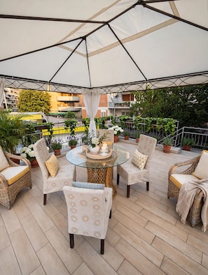 Un'area patio all'aperto con un tavolo rotondo in vetro, sedie in vimini e un tettoia bianca.