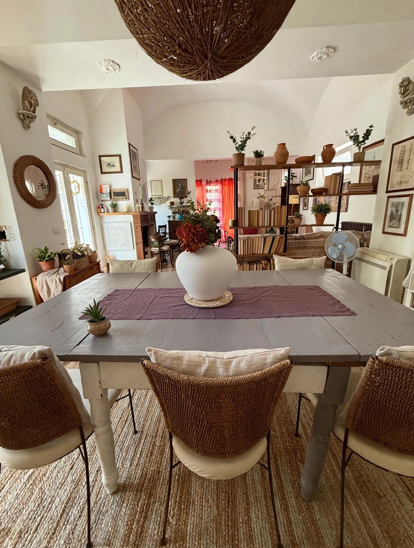 Un'area pranzo con un tavolo grande, sedie in vimini, un vaso bianco con fiori e una ventola.