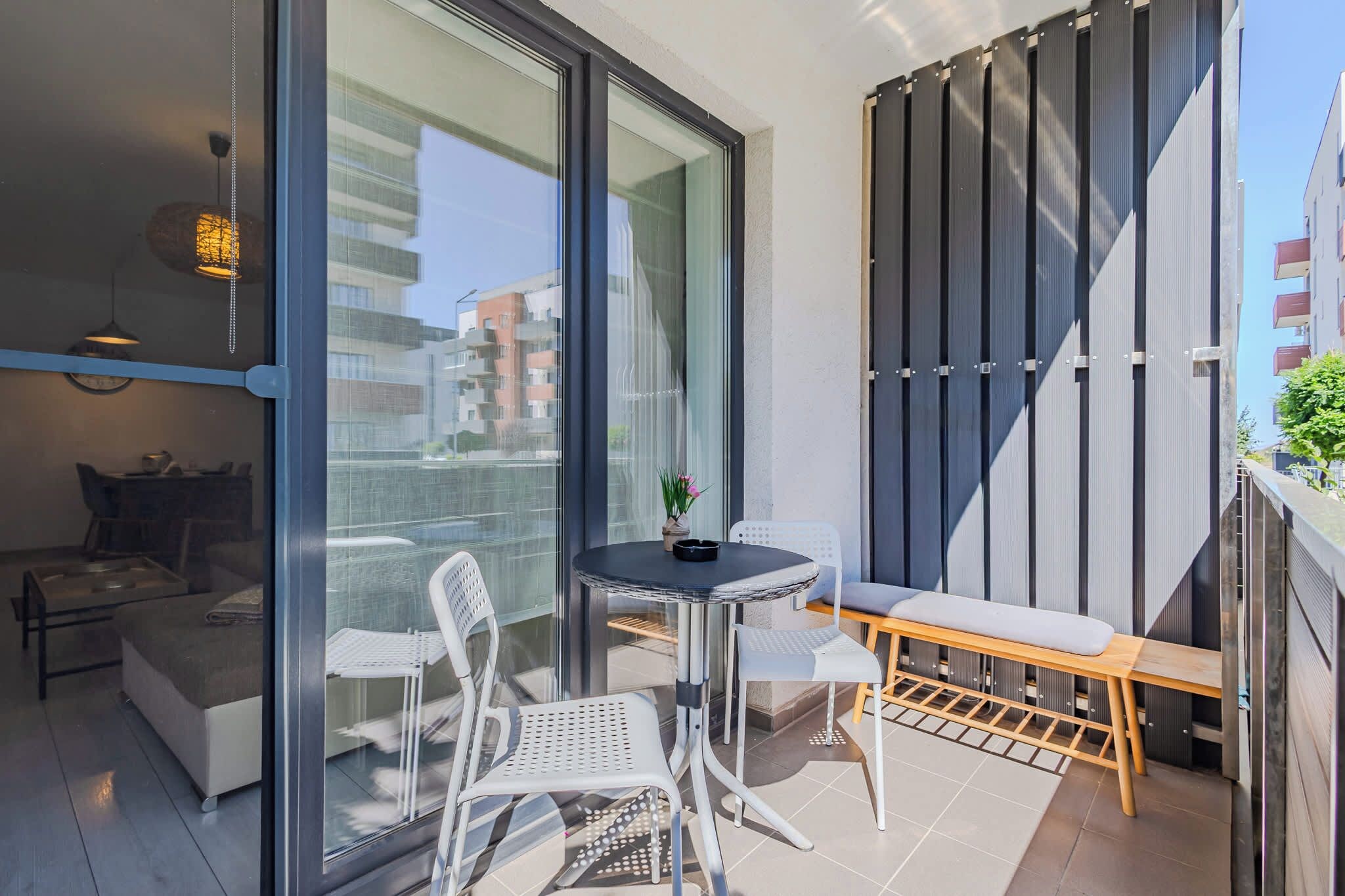 Un balcon moderne avec une porte vitrée, une table ronde, deux chaises, un banc et une plante en pot.