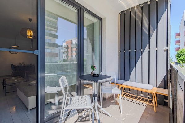Un balcón moderno con puerta de vidrio, una mesa redonda, dos sillas, un banco y una planta en maceta.