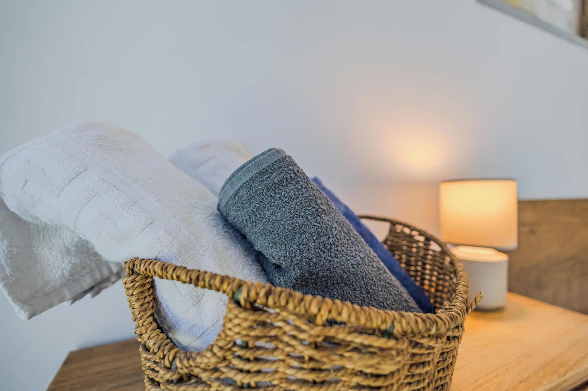 A woven basket containing folded towels on a wooden surface with a lamp in the background.
