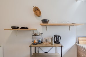 Una cocina con estantes de madera que contienen boles y vasos, una cafetera y una tabla de cortar sobre una mesa de madera.