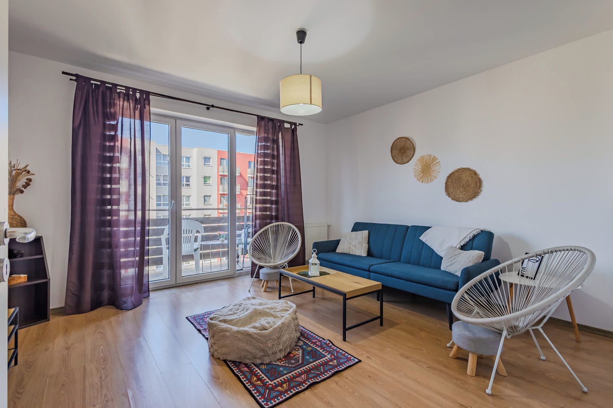 Een moderne woonkamer met een blauwe bank, twee witte stoelen van draad, een bijzettafeltje, een kamerkleed met een patroon en een groot raam met paarse gordijnen.