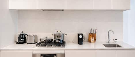 Una cocina moderna con armarios blancos, estufa a gas, cafetera, tostadora, hervidor, bloque de cuchillos y un lavamanos.