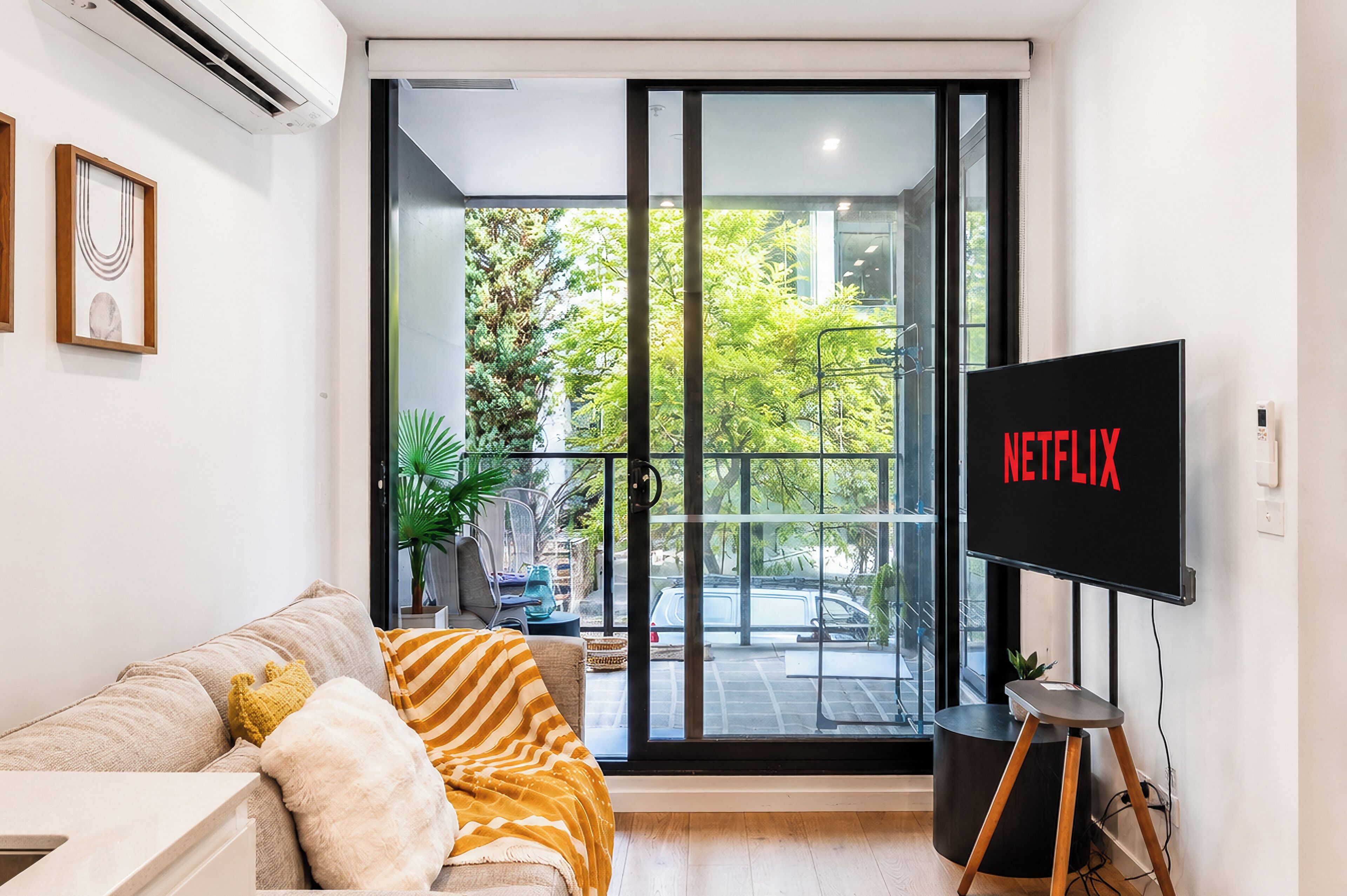 Un salon moderne doté d’une grande porte-fenêtre coulissante donnant sur un balcon, d’un téléviseur à écran plat fixé au mur et d’un canapé avec des coussins.