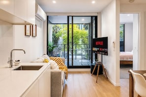 A modern kitchen with a built-in sink, a dining area with a table and chairs, a living room with a sofa and a TV displaying the Netflix logo, and a large sliding glass door leading to an outdoor area with greenery. - Hawthorn 2BR Apartment | 2 Queen Beds | Balcony (Hawthorn)