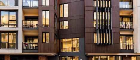A multi-story building with balconies, large windows, and a parking area with various cars.