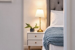 Ein ordentlich bezogenes Bett mit blauer Decke, einer Nachttischlampe und einem kleinen Topfpflanz auf einem weißen Nachttisch.