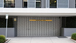 Entrada de um edifício com porta de garagem metálica, placa de restrição de altura e indicação de altura máxima de 2,1 metros.