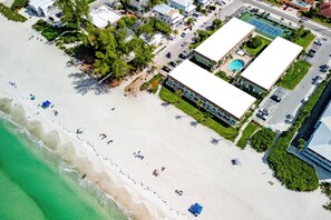 Eine Vogelperspektive auf ein Anwesen am Strand mit einem Schwimmbad, mehreren Gebäuden und einem Sandstrand mit Sonnenschirmen.