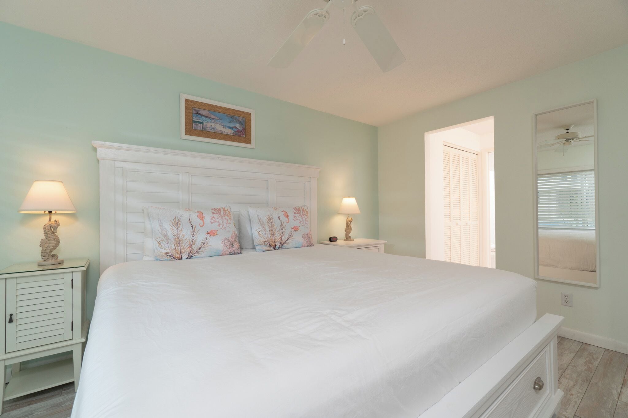A bedroom with a large bed, two bedside tables with lamps, a ceiling fan, a window with blinds, and a painting on the wall.
