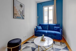 Ein Zimmer mit einem blauen Sofa, einem kleinen runden Tisch mit Kerzen, einem schwarzen Hocker und einem Bild an der Wand.