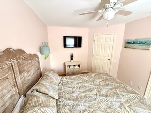 Ein Schlafzimmer mit einem Bett, einem Deckenventilator, einem Fernseher, einer Tür und einem kleinen Tisch mit einer Lampe.