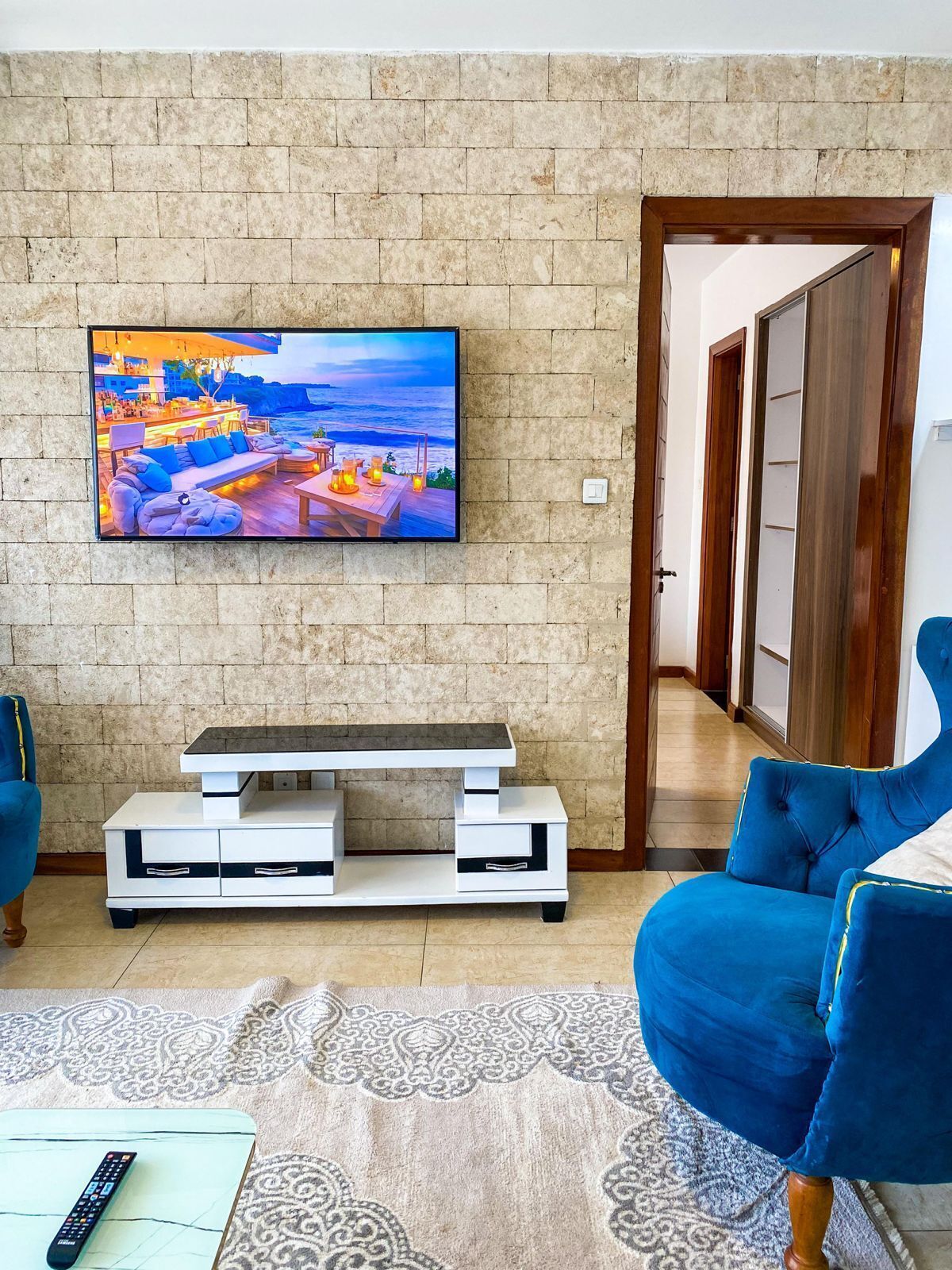 A living room with a flat-screen TV mounted on a stone wall, a white entertainment unit, a blue sofa, and a patterned rug.