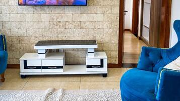 A living room with a flat-screen TV mounted on a stone wall, a white entertainment unit, a blue sofa, and a patterned rug.