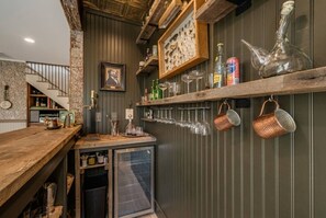 Eine Barbereich mit Holztresen, einem kleinen Kühlschrank und einem an der Wand befestigten Regal mit Flaschen und Gläsern.