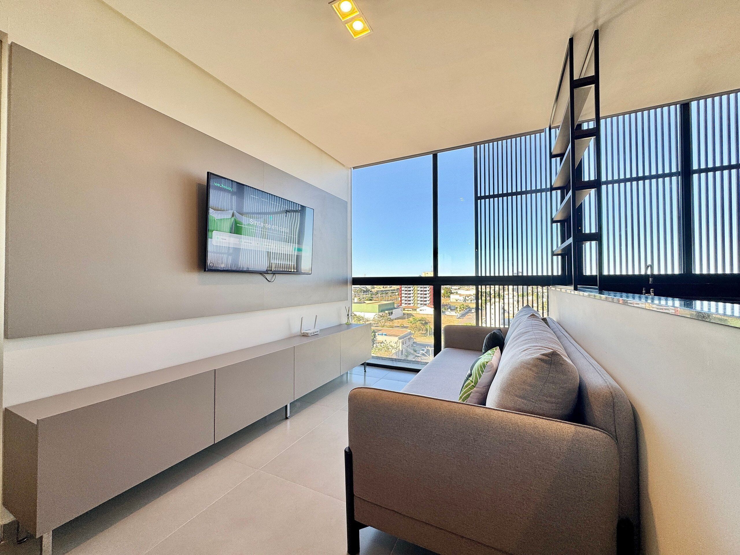 A modern living room with a sofa, a mounted TV, and a view of the city through large windows.