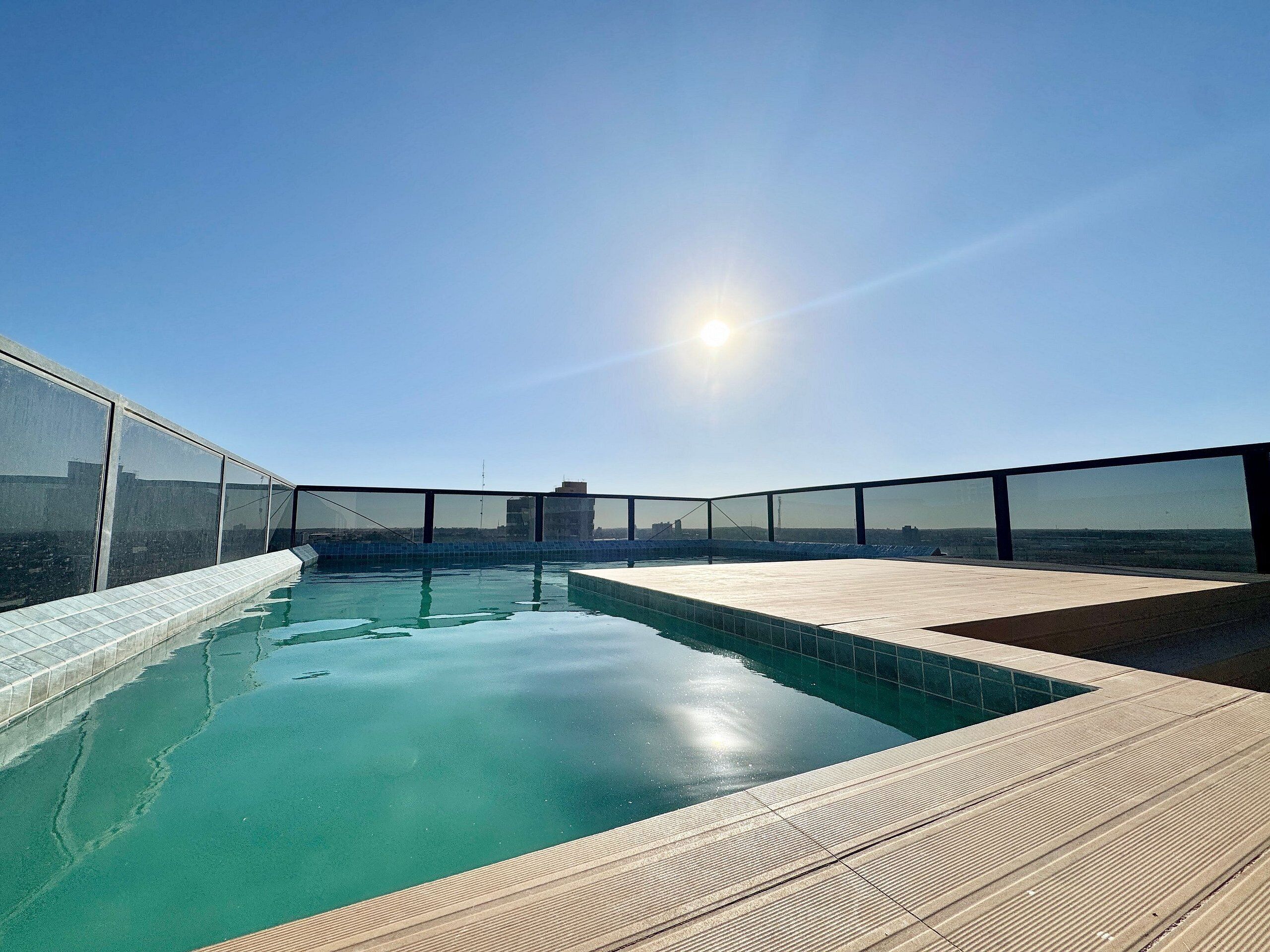 A rooftop swimming pool with clear blue water, surrounded by a wooden deck and glass railings.