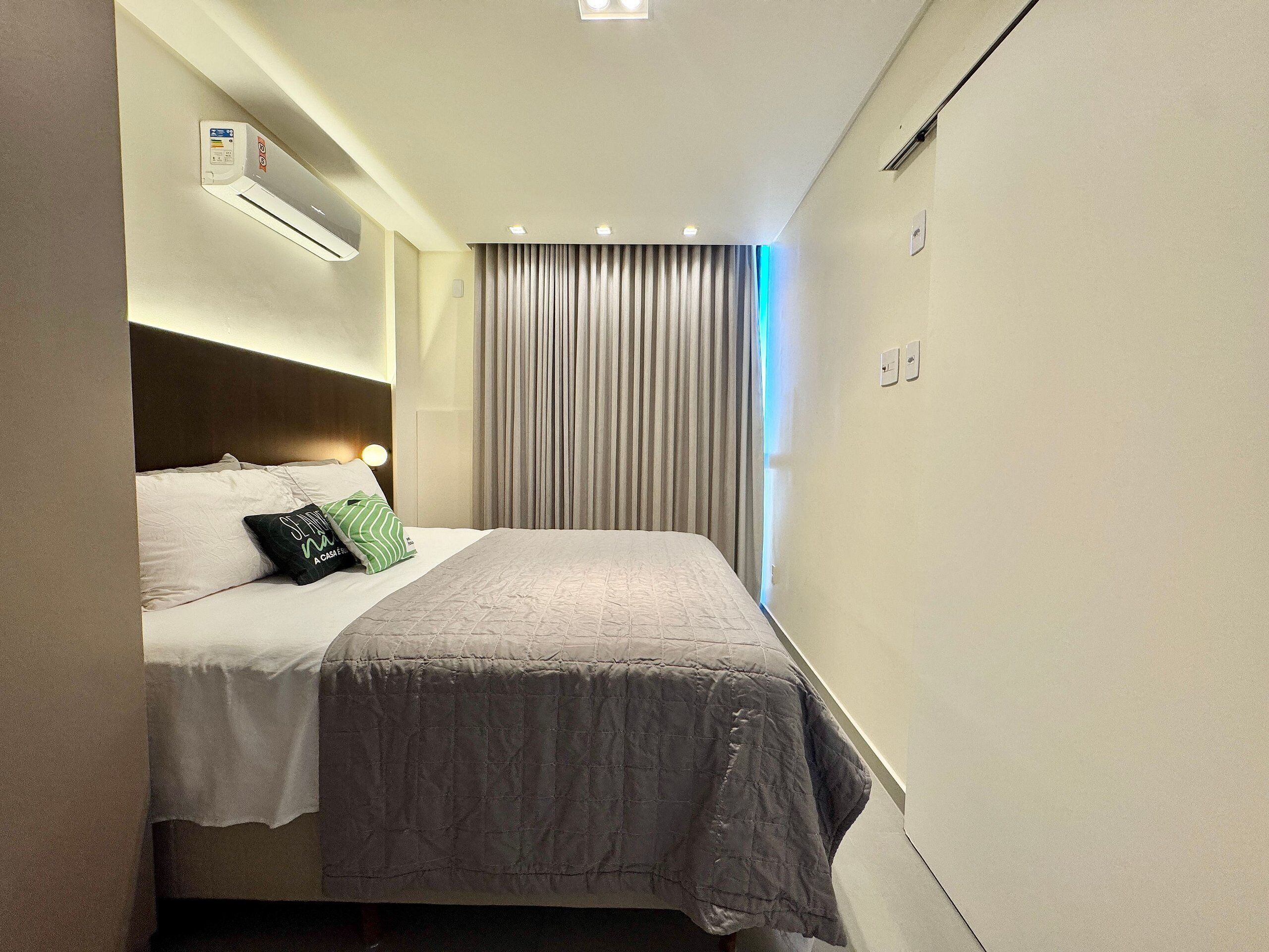 A narrow hotel room with a bed, a headboard, a window with curtains, and a wall-mounted air conditioning unit.
