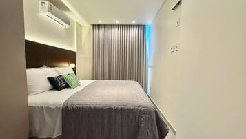 A narrow hotel room with a bed, a headboard, a window with curtains, and a wall-mounted air conditioning unit.