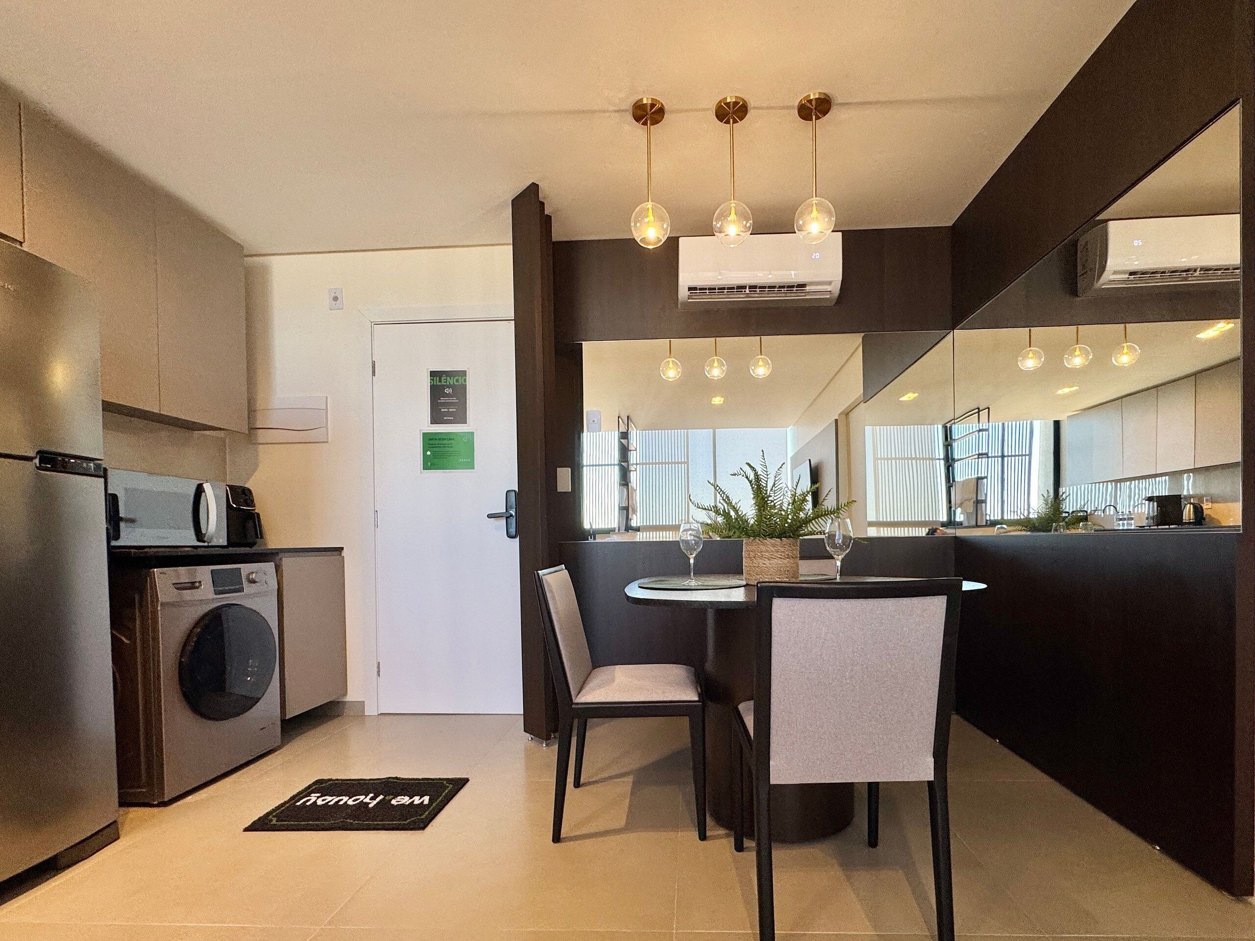 A modern kitchen with a built-in washing machine, a dining area with a round table and chairs, and a large mirror reflecting the room.
