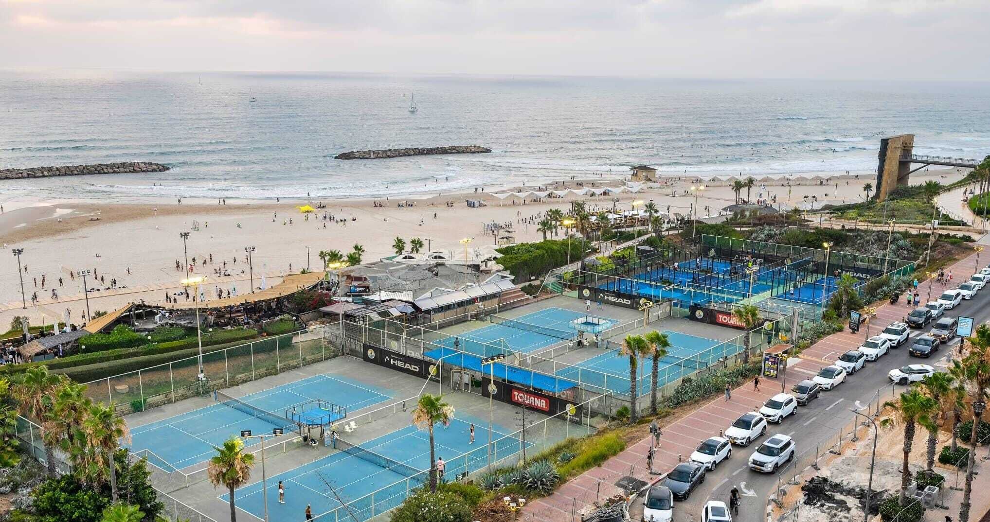 A beachside tennis court complex with multiple courts, a parking area, and a view of the ocean.