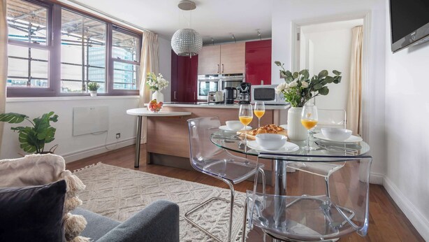 A modern kitchen with a dining area, featuring a glass table, clear chairs, and a red cabinet. - 1BR by Piccadilly Circus & Trafalgar Square (London)