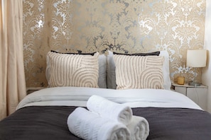 A neatly made bed with a dark headboard, two pillows with a textured pattern, and a rolled white towel on the bed. - 1BR by Piccadilly Circus & Trafalgar Square (London)