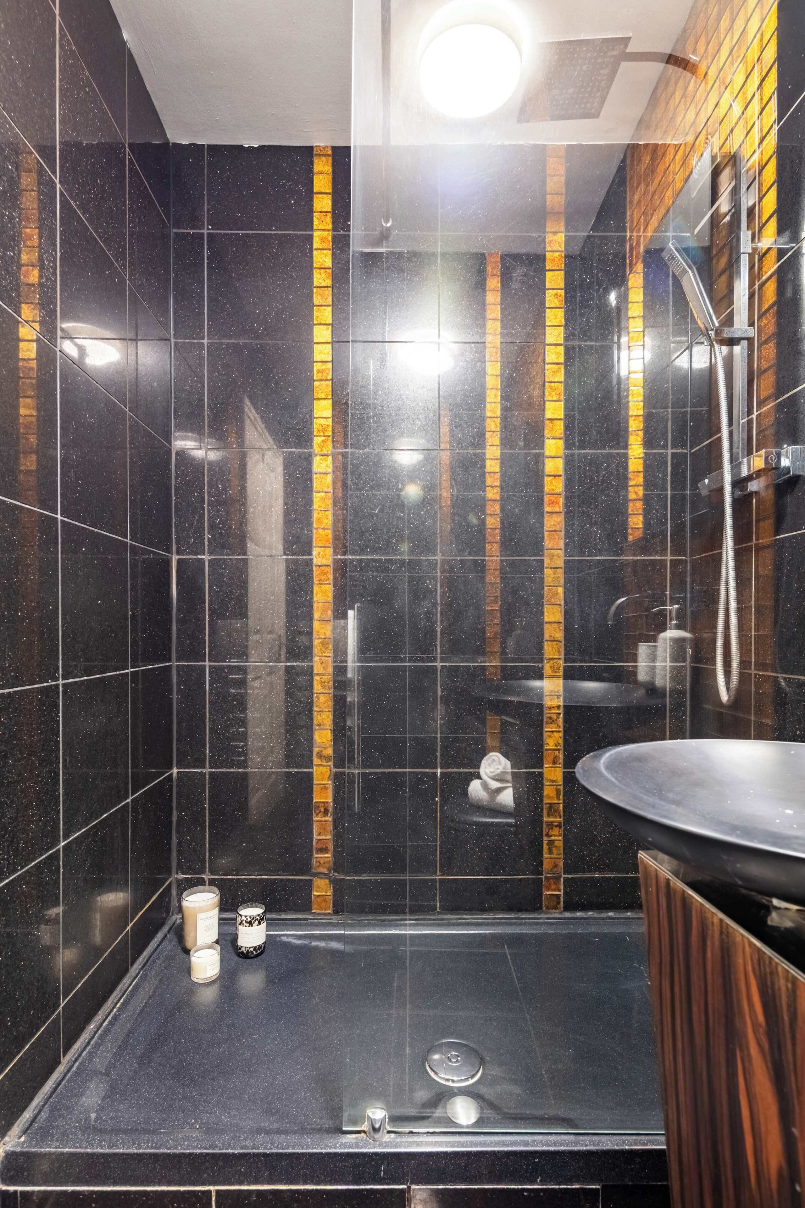 A modern bathroom with black tiled walls, a wooden accent wall, a shower area, and a sink.