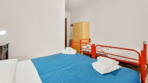 Un letto singolo con copriletto blu e cuscini bianchi, un letto a castello in metallo rosso e un armadio in legno in una stanza dalle pareti bianche.