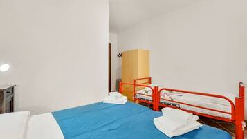 Een eenpersoonsbed met een blauwe spreideken en witte kussens, een rode metalen stapelbed en een houten garderobe in een kamer met witte muren.