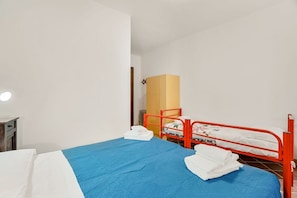 Ein Einzelbett mit blauer Bettdecke und weißen Kissen, ein rohes Metall-Etagenbett und ein Holzschrank in einem Zimmer mit weißen Wänden.