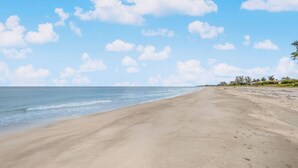 A wide sandy beach with gentle waves, clear skies, and scattered clouds.