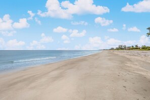 A wide sandy beach with gentle waves, clear skies, and scattered clouds.