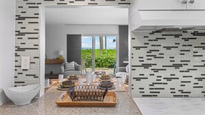 A modern kitchen with a dining area, featuring a kitchen island with a metal rack, a view through the window, and a white cabinet.