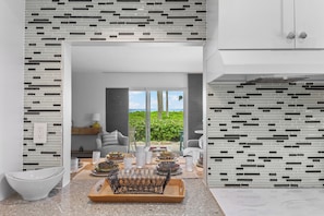 A modern kitchen with a dining area, featuring a kitchen island with a metal rack, a view through the window, and a white cabinet.
