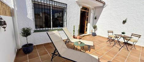 Un patio con mesa y sillas, un pequeño jardín con plantas en macetas y una ventana con rejas.