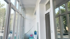 A narrow corridor with large windows on both sides, a small seating area with cushions, and a door on the right.