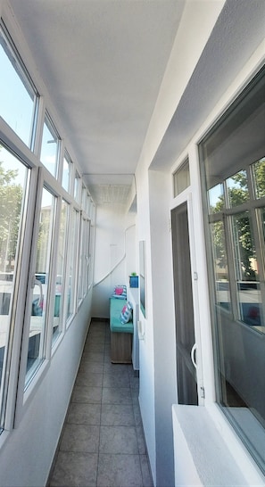 A narrow corridor with large windows on both sides, a small seating area with cushions, and a door on the right.
