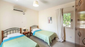 Ein Schlafzimmer mit zwei Betten, einer Klimaanlage, einem Nachttisch mit Lampe, einem gerahmten Bild an der Wand und einem Fenster mit Vorhängen.