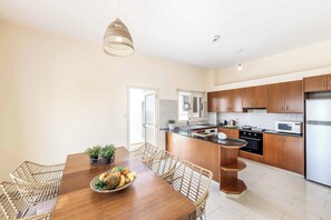 A modern kitchen with wooden cabinets, a central island, and a dining area with wicker chairs. - Coralia Dream 1 - Three Bedroom Villa, Sleeps 6 (Peyia)