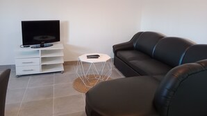 Un salon moderne avec un téléviseur à écran plat posé sur un meuble blanc, un canapé en cuir noir, une petite table d’appoint blanche avec une télécommande, et un petit tabouret blanc.