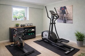 A home gym with an elliptical, a stationary bike, and a bench press. There is a window with plants on the sill, a shelf with kettlebells, and a poster on the wall. - Villa Manatee - Wellness Apartment with a Balcony (Sellin)