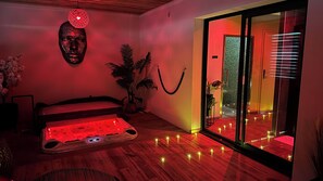 Ein Zimmer mit einem Bett, einer Massageliege, einer Wandskulptur eines Kopfes, einem Topfpflanzen und einer Schiebetür zu einem Außenbereich.