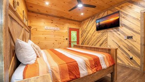 A wooden cabin room with a bed, a ceiling fan, a television, and a red door. - 🎱 Riversong Elegance | Game Room, Hot Tub & Sleeps 12, Minutes to Dollywood 🏔️ (Sevierville)